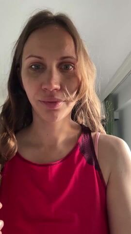The video shows a woman indoors, standing and facing the camera. She is wearing a red tank top. She has long brown hair, and she is running her hands through it. The background is a plain room.