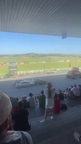 The video shows a red monster truck performing a stunt, driving over two cars. The stunt is being watched by a crowd of people sitting in bleachers. The setting appears to be an outdoor event with a racetrack and green fields in the background. The sky is blue and sunny. The monster truck is red with a white stripe and has large wheels. The cars are silver and dark gray. The people in the crowd are dressed in casual clothing. The video is filmed from the perspective of someone in the crowd, looking towards the stunt area.