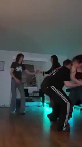 Four young adults are dancing in a room with wood floors. One person wears a black t-shirt and jeans and dances solo at the edge of the frame. A couple dances in the center, with the male wearing a black t-shirt and Adidas pants and the female wearing a red top and loose jeans. He lifts her into the air during the dance. A fourth woman wearing a black dress and black tights circles the dancing couple. A small entertainment center is in the background with turquoise lighting. The walls are white, and the ceiling is white with a light. Overall, the scene depicts friends dancing in a casual, indoor setting.