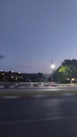 The video shows a blurry view of a gas station at dusk. There is a tree on the left side of the screen and the gas station is on the right. The gas station has red and white signage and lights are on. A cell tower is visible in the distance. The sky is a pale blue.