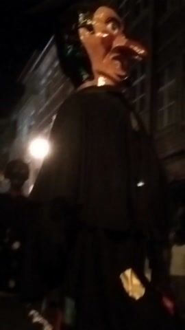 A dimly lit, outdoor scene shows a group of people interacting. One person, wearing a dark outfit and a green scarf or hat, is partially obscured by what appears to be a dark tarp or fabric. This person is holding a colorful object with red and green accents. Other figures are visible in the background, also appearing to be in dark clothing, with some indistinct objects and structures. The overall impression is that of a nighttime gathering or event, possibly a street festival or performance, with limited visibility.