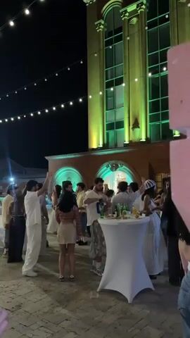 The video shows an outdoor gathering at night in front of a building. The building is illuminated with changing colored lights, starting with green, then transitioning to blue. There are string lights hanging overhead. People are dressed in casual attire and appear to be socializing. Some are standing, some are talking in groups, and some are near a white cocktail table with drinks on it. The ground is paved. The overall atmosphere appears to be a party or event.