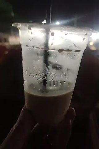 A hand holds a partially consumed beverage in a clear plastic cup with a lid. Condensation covers the exterior of the cup. The drink is a light brown color. A black straw is visible inside the cup. In the blurred background, indistinct shapes and lights suggest a nighttime outdoor environment. The speaker mentions "Jumatan Menteng."