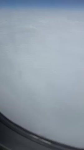 The video is a first-person view from an airplane window, looking out at a vast expanse of clouds under a clear blue sky. The clouds are white and appear to be layered, with some areas looking slightly rippled or textured. The bottom portion of the airplane window frame is visible, and the light outside suggests it is daytime. There are no people or specific objects visible within the frame apart from the clouds and the sky. The camera appears to be static, offering a consistent perspective of the aerial landscape.