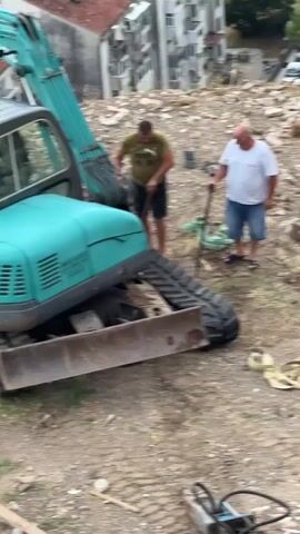 The video shows two men working near a teal excavator on a debris-filled hillside in Igalo, Montenegro. The excavator's bucket is raised, and its tracks are visible. The man on the left, wearing a green t-shirt and black shorts, is actively engaged with the excavator's track, appearing to manipulate it with a rope. He is barefoot. The man on the right, in a white t-shirt and blue denim shorts, stands nearby holding a long-handled tool, possibly a shovel or pickaxe, and observes the activity. The ground is uneven and covered in rubble, dirt, and scattered construction materials.

In the background, a multi-story building is visible, suggesting an urban or semi-urban environment undergoing demolition or renovation. Several cars are parked on a road adjacent to the hillside. The lighting suggests it is daytime, likely with clear weather conditions. The overall scene depicts manual labor and heavy machinery operation on a construction or demolition site.

Visible text on the excavator's body reads: "PRATOF PAVLOVIC | C.A.A. BRKOVIC | 2014 G. RAD | 068 006 201"