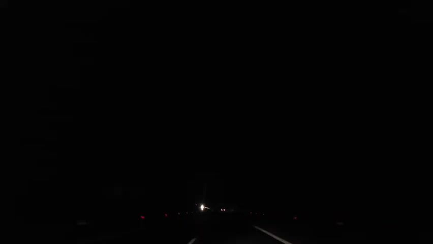 The video displays a dark, nighttime scene from a low, forward-facing perspective, likely from a moving vehicle. A faint road is visible, marked by a solid white line on the right. On the left, several small, reddish-orange lights, possibly road reflectors, are spaced out along the side of the road. In the far distance, a single bright white light, likely an oncoming vehicle's headlight, approaches slowly. No other significant objects or people are visible.