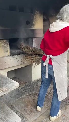 A woman wearing a red hooded sweatshirt, blue jeans, an apron, and white sneakers is shown from behind using a bundle of pine branches to clean out a brick oven.  She puts the pine branches into the dark interior of the oven, which is set into a structure made of tan-colored bricks. A wooden bench is partially visible to the left.  The woman moves the branches around inside the oven.