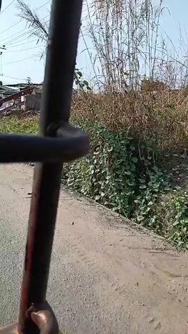 The video shows a view from a moving vehicle, focusing on a metal frame that obstructs the view. Outside, the road is paved and cracked. A pile of what appears to be playground equipment, including a slide, is visible on a dirt patch next to the road. There is a white car parked near the playground equipment. Buildings are visible in the background, along with power lines running overhead. The sky is clear and bright.