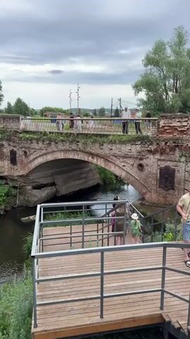 The video shows an old brick bridge spanning a river. Several people are visible on the bridge and on a wooden walkway leading to the bridge. The bridge has an arched design and appears to be in a state of disrepair, with some bricks missing and vegetation growing on it. A plaque on the bridge shows the year 1855. The sky is cloudy, suggesting overcast weather. The surrounding environment appears to be natural, with trees and greenery visible.