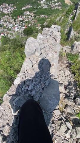 The video shows a person standing on a stone structure, overlooking a town nestled in a valley surrounded by mountains. The scene is captured on a sunny day with scattered clouds in the sky. The stone structure appears to be part of ruins, with rough, gray stones stacked together. The town below consists of buildings with red roofs, interspersed with greenery. The surrounding mountains are covered in dense forests. A road winds through the valley, connecting the town to other areas. The camera slowly pans, revealing more of the ruins and the landscape. A quarry or industrial site is visible in the distance.