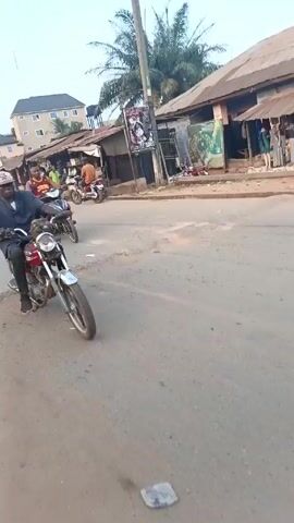 The video shows a street scene in what appears to be a small town or village. A man wearing a hat and a dark shirt is riding a red and chrome motorcycle. Several other motorcycles pass by. People are walking on the side of the road near buildings that appear to be small shops or stalls. The road surface is paved, but shows signs of wear and tear. A few larger buildings are visible in the background. The time of day seems to be early morning or late afternoon, as the light is somewhat muted.