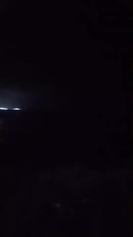 The video begins in near-total darkness, with only a faint light visible in the upper left corner, possibly from distant buildings or streetlights. As the video progresses, the camera pans slightly, revealing a dark, unlit landscape that appears to be rural or undeveloped. The lower portion of the screen shows dark, indistinct shapes that could be vegetation or ground cover. The ambient light source remains the same and does not change throughout the brief recording. There are no discernible people, animals, or specific objects within the frame. The environment is entirely dark, suggesting it is nighttime or the video was captured in a poorly lit area. The overall impression is one of extreme low light conditions.