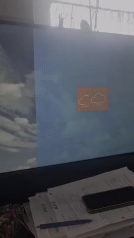 The video shows a computer monitor displaying a weather forecast. In the first few seconds, a cloud with raindrops is visible on a blue, cloudy background. This then transitions to a screen with a turbine on a windy day, with waves on the left side and a cloud icon. Finally, the screen shows a sun icon on a light gray background with an orange vertical stripe. Below the monitor, a stack of papers, a pen, and a smartphone are visible.