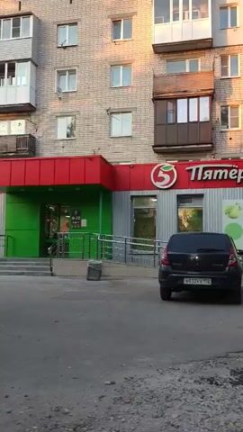 A stationary exterior shot captures a street scene in Bor, Russia. The camera is focused on a "Pyaterochka" supermarket, a well-known Russian grocery chain, with its prominent red and green signage. A dark colored car is parked in front of the supermarket, obscuring part of its entrance. To the right of the supermarket, a gold colored SUV is visible. Further down the street, a silver sedan is parked. The buildings in the background are typical of Soviet-era apartment blocks, with multiple stories and rows of windows. Trees with green foliage line the street, suggesting it is either spring or summer. The sky is a pale blue, with soft light suggesting it's either early morning or late afternoon. The ground in front of the supermarket is paved with asphalt that appears to be in disrepair, with visible potholes. The overall impression is of a typical urban landscape in a Russian town.