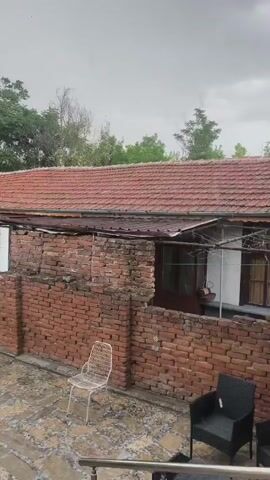 The media file captures an outdoor scene with a focus on a weathered brick building and its surroundings. A reddish-brown tiled roof covers the structure, which appears to be a house or a similar residential building. The brickwork is aged and somewhat dilapidated, with visible signs of wear and tear.

In the foreground, there are two chairs placed on a tiled patio. One is a light-colored, wire-frame chair, and the other is a darker, wicker-style armchair. The patio itself has a textured, stone-like surface.

Behind the building, lush green trees are visible, suggesting a natural or semi-rural setting. The sky above is overcast and gray, indicating cloudy weather. There are no people visible in the frame.

The overall impression is one of a quiet, possibly abandoned or infrequently used outdoor space. The weather appears to be damp, perhaps due to rain or recent rainfall, as suggested by the gray sky and potentially wet surfaces. The scene evokes a sense of stillness and perhaps a touch of melancholy due to the aged appearance of the building and the somber sky.

The location context is Harmanli, Bulgaria. There is no visible text within the media file.