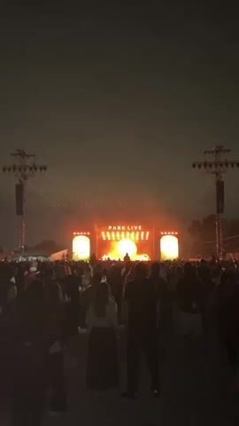 A large crowd of people stand facing a stage at an outdoor concert at night. The stage is lit with pink and orange lights, and the words "PARK LIVE" are displayed in large letters at the top of the stage. Two tall speaker towers are positioned on either side of the stage, and bright spotlights illuminate the sky. The sky is dark and hazy, suggesting it might be cloudy or filled with smoke. Most of the people in the foreground are silhouetted against the lights of the stage, making their features indistinguishable. One person in the center foreground is wearing a long, dark skirt and a lighter top. Another person in the foreground is wearing a floral patterned shirt. The overall atmosphere appears to be that of a lively music festival.