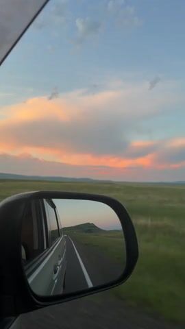The video shows a view out of a car window, with a side mirror reflecting the road behind. The sky is a mixture of blue with scattered clouds tinged with pink and orange from a sunset. The landscape outside the car consists of green fields and rolling hills. The road is paved and appears to be a highway. The car is moving, as indicated by the blur of the passing scenery. The side mirror shows the car's own reflection, the road stretching back, and a distant hill. No people are visible in the frame.