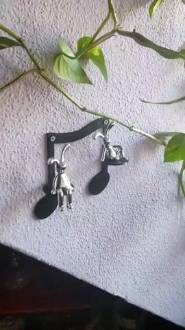 The video shows a series of indoor plant arrangements. The first scene shows a green and yellow vine-like plant hanging on a textured, light gray wall. Two black musical note-shaped hooks with metallic rabbit figurines are attached to the wall, seemingly supporting the plant. The second scene focuses on a tiered plant display with metal buckets containing various plants. A ceramic squirrel figurine sits amongst the greenery. The final scene displays a plant in a red pot, placed next to a window. Two tall, giraffe-shaped decorations made of natural materials are positioned behind the plant, along with a small white mushroom decoration in the soil.