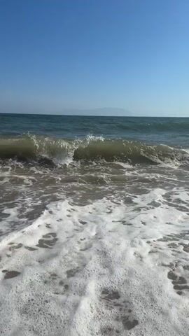 A short video captures ocean waves breaking on a sandy shore under a clear blue sky in Νέα Χηλή, Greece. The video begins with a wave rolling in and breaking, creating a significant amount of white foam that spreads across the sand. The foamy water then recedes, revealing the wet sand. Another wave can be seen breaking further out in the ocean. The horizon features a faint line of distant land or mountains. The sunlight is bright, suggesting a clear, sunny day. The scene is devoid of people or visible objects other than the natural elements. The natural sounds of the waves crashing are audible throughout the clip.