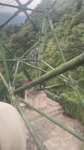 The video begins with a close-up of a large green metal structure, possibly part of a cable car support tower, with diagonal beams and circular connectors visible. Several people are gathered on a concrete platform at the base of the structure. One person in a red shirt stands on the platform, and another person in a white helmet is also visible. Above, on the tower, a group of people are seen in a suspended platform or cage, with ladders extending upwards. The camera then pans to show a dense, green, forested mountainside. The perspective shifts to look through a window, revealing a cable car in the distance moving across the sky above the trees. The final shot focuses on the back of a person's head wearing a white mesh baseball cap, with the cable car and the misty, forested mountains visible through the window. The overall scene depicts a cable car ride through a mountainous, wooded area, with glimpses of infrastructure and other passengers.