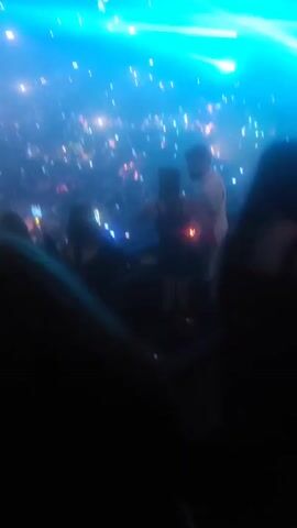 The video shows a large crowd of people at a concert or performance. The crowd is dense, with many people holding up their phones, presumably recording or taking pictures. Above the crowd, there is a light show with bright beams of light in various colors including blue and green. The environment appears to be a large indoor venue, possibly a stadium or concert hall. The overall atmosphere is energetic and festive.