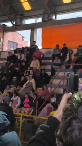 The video shows a crowded indoor arena with people sitting in bleacher seating. The seating is mostly filled with a diverse group of people of all ages, some wearing traditional clothing. A brown metal support column obstructs some of the view of the seating. Behind the seating is a large orange wall. The environment appears to be a community event, possibly a sporting event or cultural gathering, as suggested by the bleacher seating and crowd. The video is shaky and the camera moves around, suggesting it's handheld.