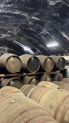 The video shows the inside of a distillery warehouse full of barrels. The barrels are stacked on top of each other in rows, and they appear to be made of wood. The room is dimly lit, with just a few overhead lights. The walls are made of brick and stone, and the ceiling is covered in mold. The camera pans across the room, showing more of the barrels and the warehouse. At the end, it shows a door leading into the warehouse with the words "TULLIBARDINE DISTILLERY WH No. 4 BLACKFORD" printed on it. Through the open gate next to the door, there are more barrels visible in the background. Above the gate, there are the words "STILL WHISPERING AND ANGELS AT WORK".