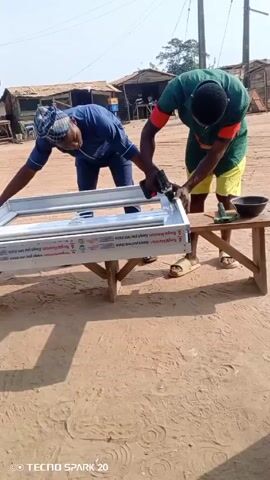 Two men are working on a white aluminum window frame. They are outside, standing on a sandy surface next to a makeshift wooden table. The man on the left is wearing a blue shirt, dark pants, and a patterned hat. The man on the right is wearing a green and orange shirt and yellow shorts. He is using a power drill to assemble the frame. There is a small black bowl and a can of lubricant on the table. The background shows simple buildings and utility poles under a clear sky. A “TECNO SPARK 20” watermark is visible in the lower left corner of the frame.