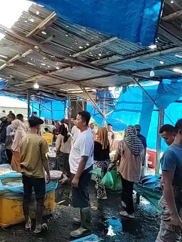 The video depicts a bustling fish market under a corrugated metal roof and a blue tarp. Several people are walking around, some wearing hijabs or face masks. There are containers of fish visible, and the floor appears to be wet. The atmosphere suggests a busy commercial environment.