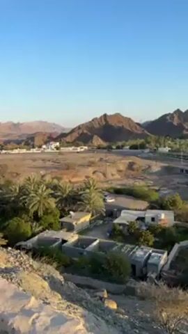 The video begins with a distant view of brown mountains under a clear blue sky, then slowly pans right to reveal a small town nestled among the trees and mountains. A small stone structure sits in the foreground. As the camera continues its rightward pan, a row of houses is visible, and the sun begins to emerge over the mountains, its light creating a lens flare. The video concludes with a view of the mountains illuminated by the sun, the town situated at their base.