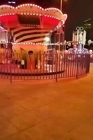 The video shows a carousel ride at night. The carousel is lit up with colorful lights that change. The ride appears to be empty, with no people visible on it. The surrounding area is paved and there are some fences around the ride. In the background, there are tall buildings and other amusement park rides, also illuminated with lights. The lights on the carousel are primarily green, blue, and red, and they transition between these colors. The carousel itself has a striped pattern on its central column and a scalloped top. The overall impression is of a lively amusement park at night, though the ride itself is currently inactive.