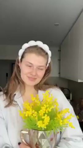 Here's a description of the video:

A young woman stands in a kitchen holding a silver vase filled with artificial yellow mimosa flowers. She is smiling slightly at the camera. She wears a white, cloud-shaped headband and a light-colored, button-down shirt. The kitchen has white cabinets and a window providing natural light. The camera is positioned at a slightly elevated angle. There is audio present with a woman speaking in Russian.