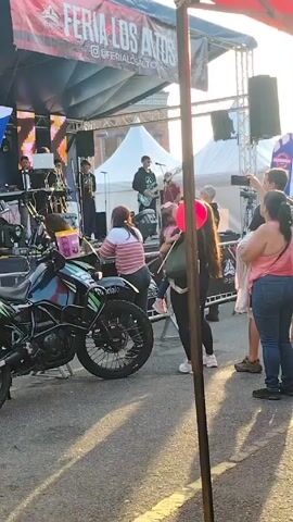 The video shows a live band performing on a stage at what appears to be a festival or fair, based on the "FERIA LOS ALTOS" banner above the stage. The band consists of at least four members playing keyboard, brass instruments, and guitars. A crowd of people is gathered in front of the stage, some passing by, some standing and listening. In the foreground, a black and green motorcycle is parked, with a woman standing next to it. She appears to be interacting with someone near the bike. A girl with a red balloon is also visible in the crowd. The scene is set outdoors, with tents and structures suggesting a fair or festival ground. The lighting indicates it's daytime, possibly late afternoon.