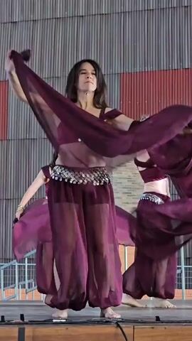 A group of women in matching purple belly dancing outfits are performing on a stage. The outfits consist of cropped tops adorned with silver sequins, flowing sheer harem pants, and coin-embellished belts. Each woman has a long, sheer purple scarf, which they are manipulating with their arms. The woman in the foreground is in the center of the frame, facing right, with her arms extended and scarves flowing. Two other women are visible behind her, also engaged in the dance. The stage is wooden and appears to be outdoors, with a striped wooden wall as the backdrop and metal railings to the left. The lighting suggests it is daytime, possibly late afternoon.