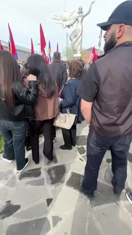 A crowd of people stands in a paved area, possibly a town square, facing a tall silver statue of a woman holding a sun-like object. Red flags, along with the Armenian flag, are interspersed throughout the crowd. The people vary in age and dress, and seem to be observing the statue. The sky is overcast. The statue appears to be a central feature of the scene, and the crowd's attention is directed towards it.