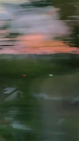 The video shows a fire on the roof of a house with solar panels. The sky is a mix of orange, pink, and yellow, suggesting a sunset or sunrise. There are trees and other houses visible in the background. The camera appears to be moving, creating a blurred effect in some parts of the video.