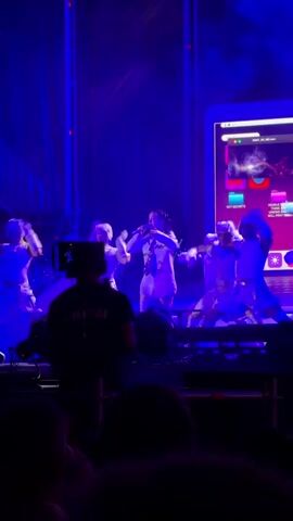 This is a video of a live performance. The stage is lit with blue lights. A female singer is on stage in a white outfit and she is holding a microphone and singing. She has dark hair pulled back in a ponytail. There are dancers on stage wearing white outfits and they are moving around the singer. There is a camera operator in the foreground filming the performance. The camera operator is wearing a black t-shirt with "CREW" written on the back. In the background, there is a large screen with some colorful graphics on it. The audience is visible in the foreground, silhouetted against the stage lights.