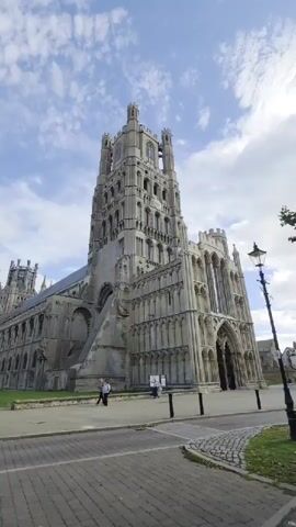 The video shows a wide shot of a large, stone cathedral with a tall tower. In the foreground, a street paved with cobblestones leads towards the cathedral. A black car drives past on the street. To the right, a lamppost stands on a grassy area. Two people are walking on the sidewalk in front of the cathedral. The sky is cloudy.