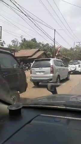 The video shows a view from inside a car looking out at a traffic scene. The car is stopped on a road, likely in a rural or suburban area given the presence of trees, power lines, and simple buildings. In front of the car, a silver SUV is visible, followed by a white SUV with a spare tire mounted on the back. Further ahead, other vehicles, including a black SUV and a red car, are parked or moving. Some motorcycles are also visible. A flag, likely red and white, is hanging from a pole. The road appears to be paved, but there are areas of dirt or gravel on the side. The sky is bright and appears to be a clear day. The overall scene suggests a moderate amount of traffic and activity.
