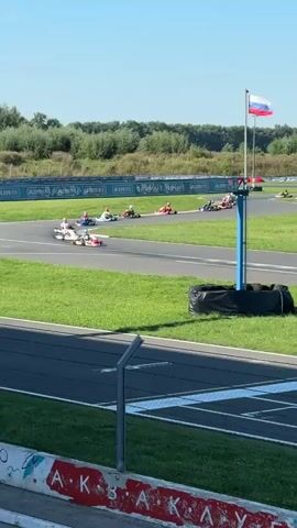 The video captures a go-kart race on a sunny day. Several karts, each with a single driver wearing a helmet, are seen navigating a winding track with green grass verges. The track has asphalt sections with white and blue markings. In the background, trees and buildings are visible under a clear blue sky. A Russian flag flies from a pole near the track. The karts are of various colors, including white, blue, pink, and black, some with sponsor logos. The drivers are positioned low in their karts. The camera perspective is from a raised vantage point, looking down at the track.