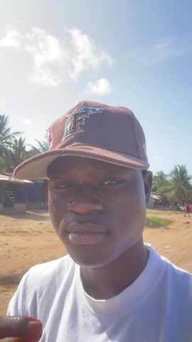 A person wearing a brown baseball cap with a Miami Marlins logo on it is filming a selfie video. They are in what appears to be a rural area with a sandy road. In the background there are buildings, trees, and some animals are grazing. The sky is blue with scattered clouds. The person holds their cap as they move. The camera remains focused on their face.