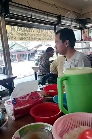 A man with short dark hair and a light grey t-shirt is sitting at a table in what appears to be an open-air restaurant. He is looking to his left. In the foreground, there are several bowls, some containing rice and what appears to be chili sauce. A large green thermal carafe is also visible on the table. In the background, through a large window or open doorway, other patrons are visible, as well as tables and chairs. A sign with Indonesian text is displayed on the window. Outside, there are vehicles and buildings.