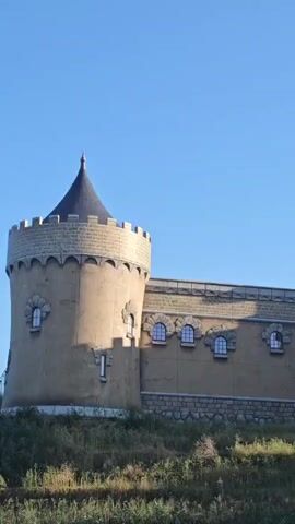 The video shows a panning shot of a castle-like structure set against a clear blue sky. The castle has beige walls, conical roofs, and small arched windows. There are also decorative battlements along the top of the walls. The castle is situated on a grassy hill. In the center of the castle facade, a sign reads "BOMBEDWORLD". The video captures the exterior of the structure as the camera moves from left to right.