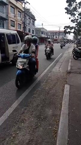 A street scene shows a two-lane road with multiple motorcycles and scooters traveling in the same direction. Buildings with multiple floors line the left side of the road, featuring windows and signs. The road is paved and has a white line dividing the lanes. The right side of the road has a sidewalk with a concrete curb. Trees are visible on the right side of the frame. The sky is overcast. In the foreground, a motorcycle with two riders passes by. Other motorcycles and scooters are seen further down the road. A white van is partially visible on the far left of the frame. The overall atmosphere appears to be a typical day in a town or city.