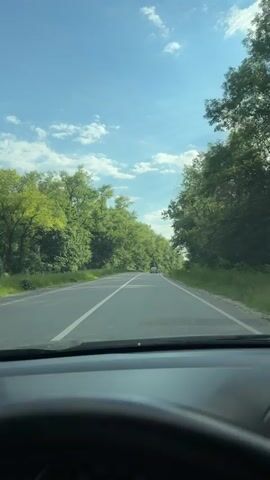 This is a first-person perspective video of driving on a paved road flanked by dense green trees. The road has a white dividing line. The sky is blue with scattered white clouds. A white car is visible in the distance, driving in the same direction. The sun is shining, casting shadows on the road. The video appears to be recorded from inside a vehicle, with the dashboard and steering wheel partially visible in the foreground. The overall atmosphere is serene and summery.