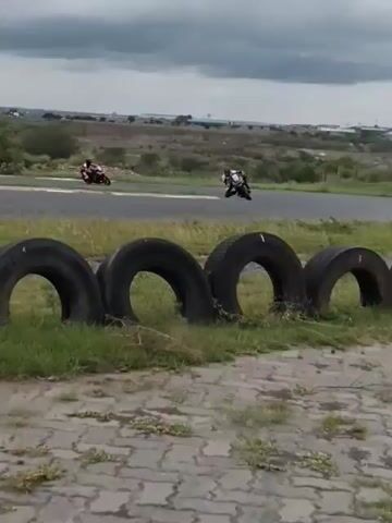 The video shows two motorcyclists racing on a paved track, framed by a barrier of old tires. The sky is overcast, and the surrounding landscape consists of sparse vegetation and distant buildings. As the motorcyclists accelerate, one pulls ahead, while the other appears to falter. The camera follows the lead motorcyclist, revealing a Coca-Cola banner in the background and more of the urban skyline.