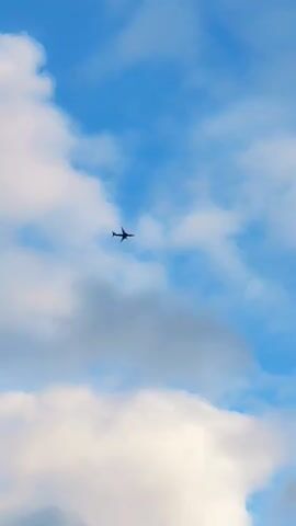 A silhouette of an airplane is seen flying against a bright blue sky with scattered white clouds. The airplane is small in the frame and appears to be moving from left to right across the sky. The clouds are fluffy and white, some are larger and denser, while others are wispy. The lighting suggests it is daytime.