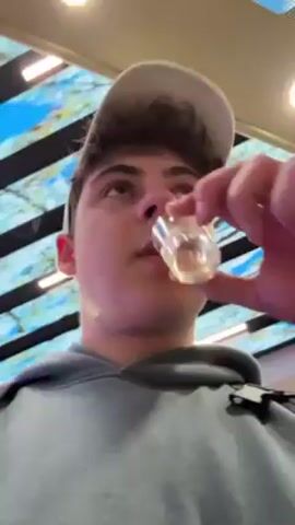 A young man in a light blue hoodie and baseball cap is drinking from a small plastic cup. He tilts his head back and another hand helps position the cup. The camera is a low-angle shot looking up at the man. The background features a modern ceiling design with linear black elements and a panel that appears to display a blue, cloud-like image.