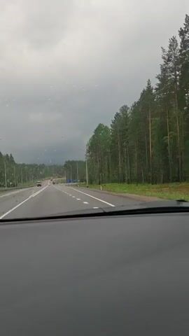The video, recorded from inside a vehicle, shows a two-lane highway with trees lining the right side. The sky is overcast and appears to be raining. The road is wet, and there are white lane markers. Several cars are visible in the distance, traveling in the same direction. One car is in the left lane, and two vehicles are in the right lane, with one appearing to be a truck. There are streetlights along the highway. A blue road sign is visible on the right side of the road. The dashboard of the vehicle is visible in the foreground.