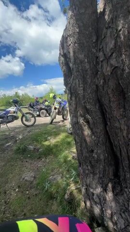 The video opens with a close-up of a large tree trunk, partially obscuring the view. As the camera pans to the right, it reveals a group of off-road vehicles, including dirt bikes and ATVs, parked on a grassy area. The scene then shifts to a breathtaking panoramic view of a lush, mountainous landscape under a partly cloudy sky. In the foreground, a person is seated on the ground, facing the expansive vista, with a dirt bike positioned to their left and gear, including a helmet and backpack, placed beside them. The camera continues to capture the serene moment of contemplation, with the rider seemingly enjoying the view. The video concludes by showcasing the same majestic scenery, with the rider still present in the frame, enjoying the tranquility of the mountaintop.